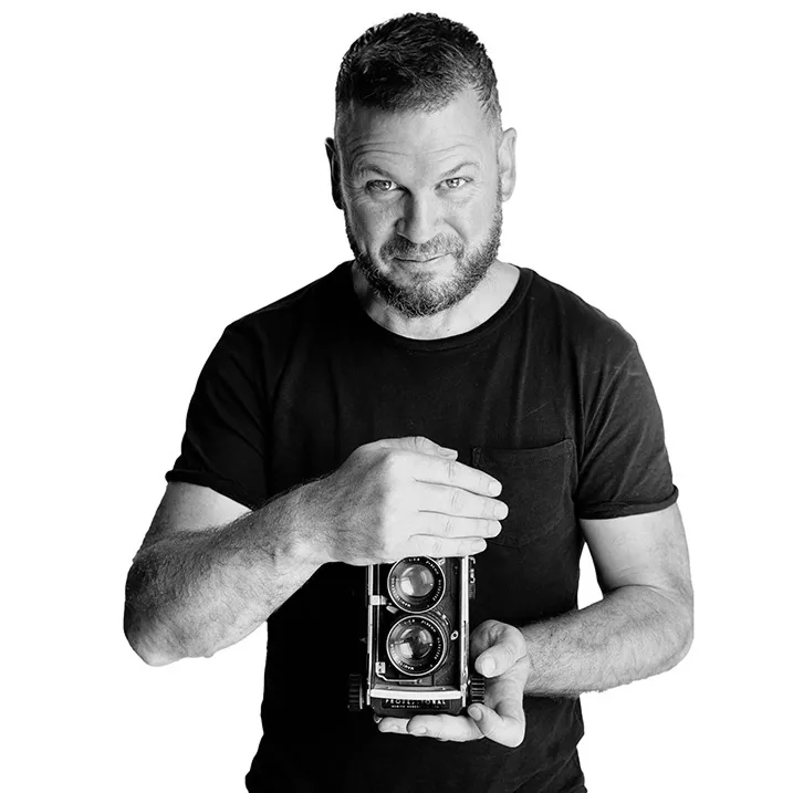 Black-and-white portrait of photographer Kevin Mark Pass in a black T-shirt holding a twin-lens camera