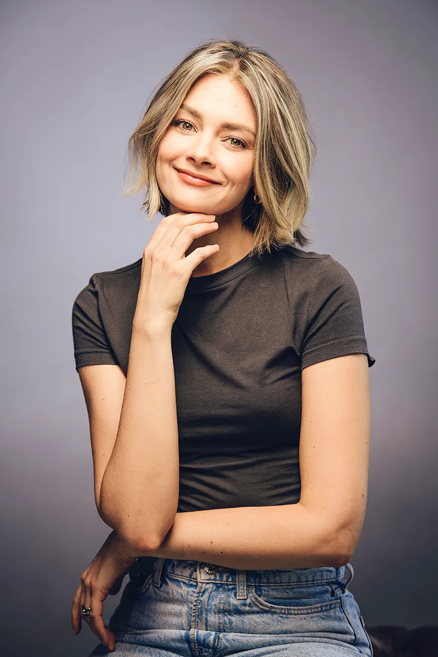 Woman with a blonde bob in a brown T-shirt smiling with chin on hand on a lilac backdrop, soft makeup look